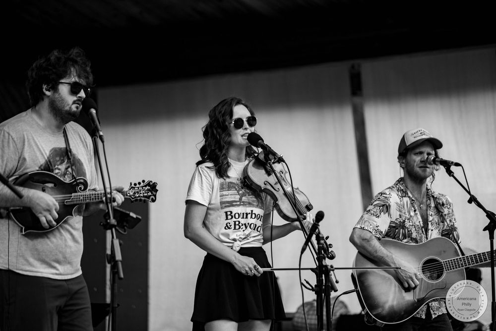 Three musicians perform on stage, two with guitars and a woman singing into a mic wearing a Bourbon & Beyond shirt.