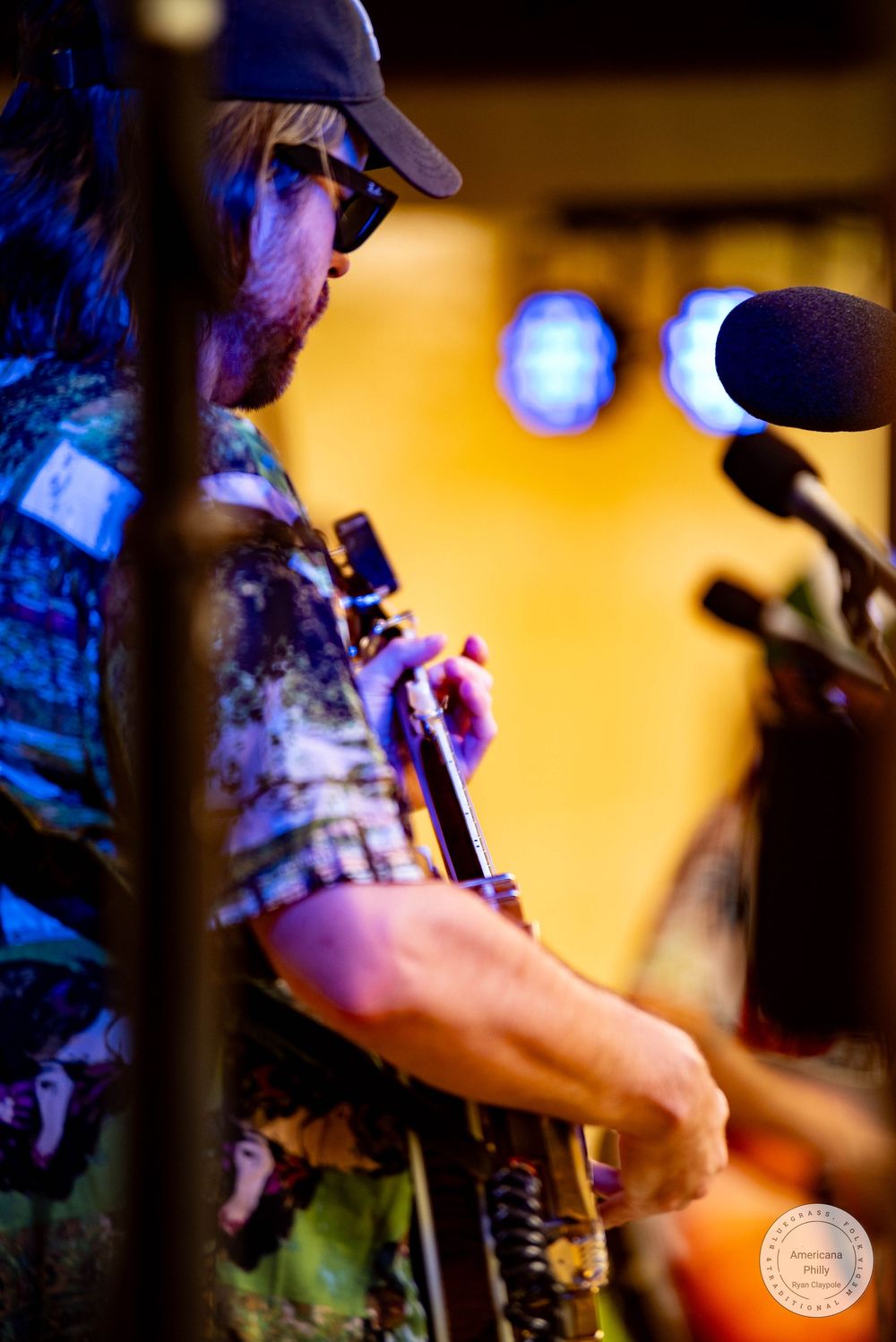 Bearded musician in a cap and colorful shirt plays guitar on stage under warm lights with a microphone nearby.