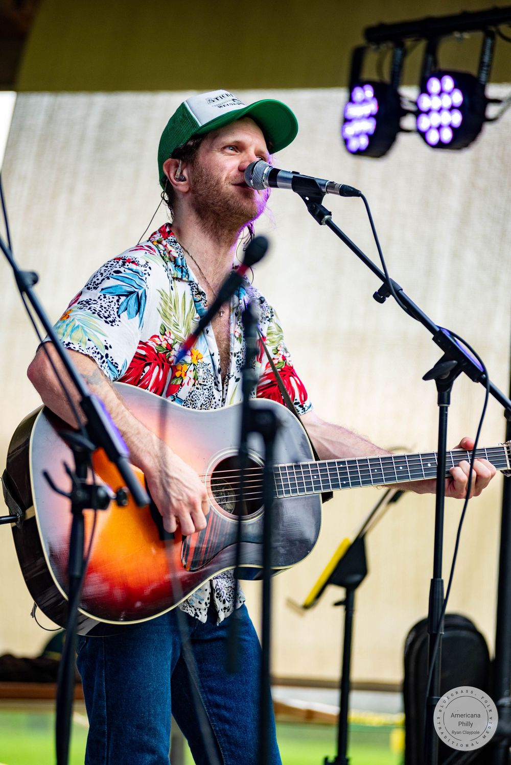 Bearded man in floral shirt and green cap playing acoustic guitar and singing into microphone on outdoor stage.