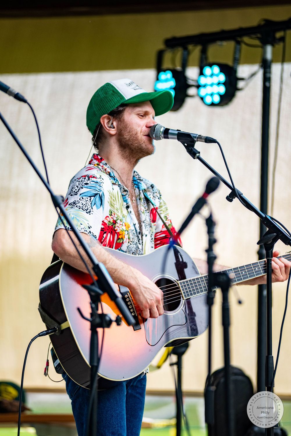 Man in green cap and floral shirt playing acoustic guitar and singing into microphone on outdoor stage.