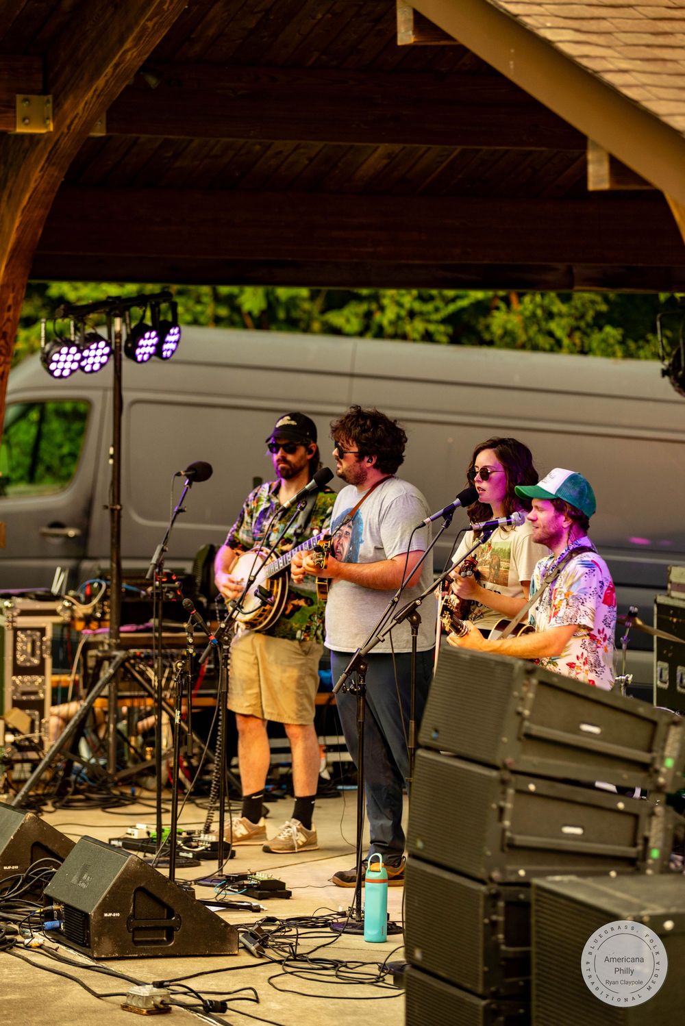Four musicians perform on an outdoor stage under a wooden pavilion, playing string instruments at a summer music festival.