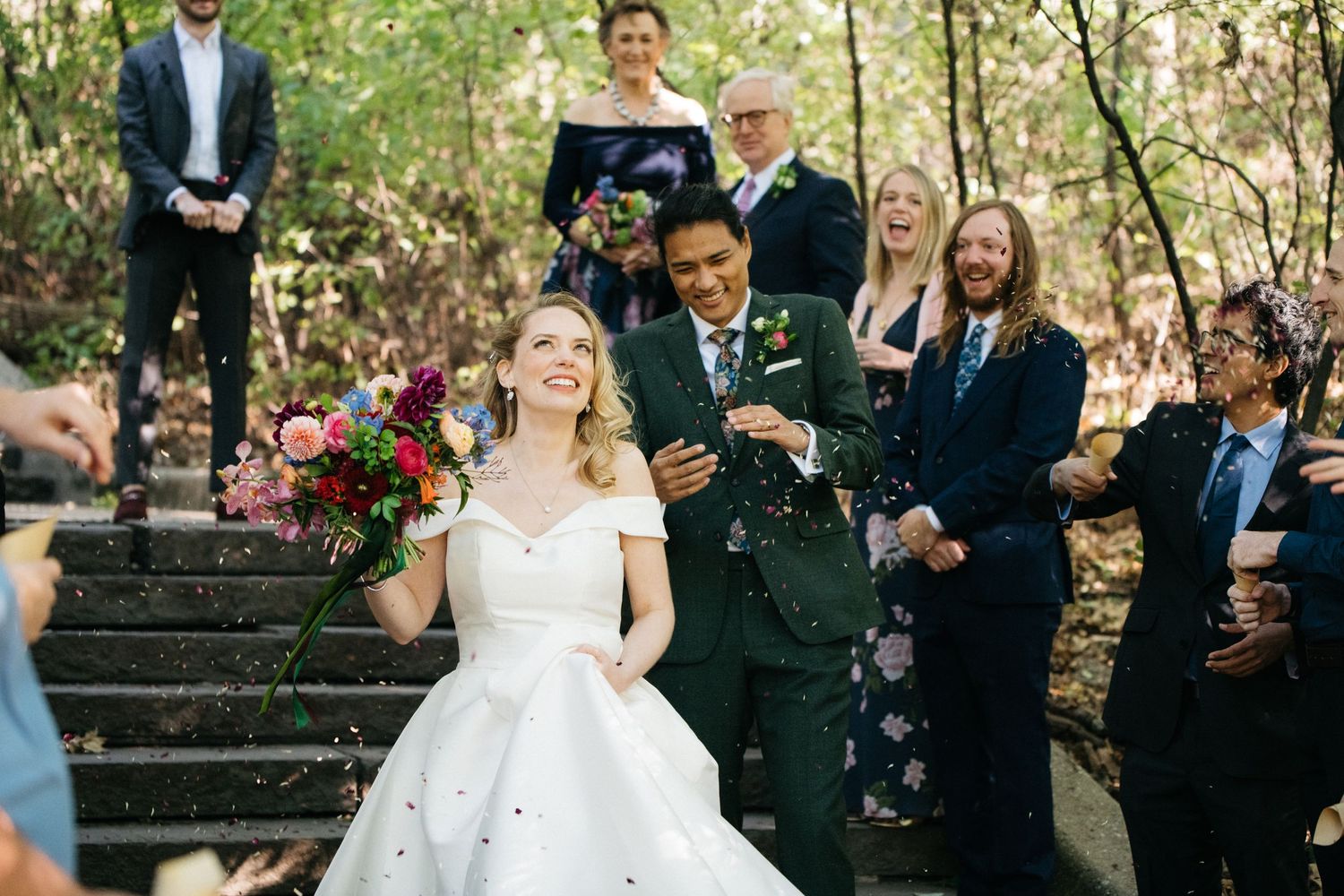 Wedding guests throw confetti as newlyweds celebrate on outdoor stone steps.