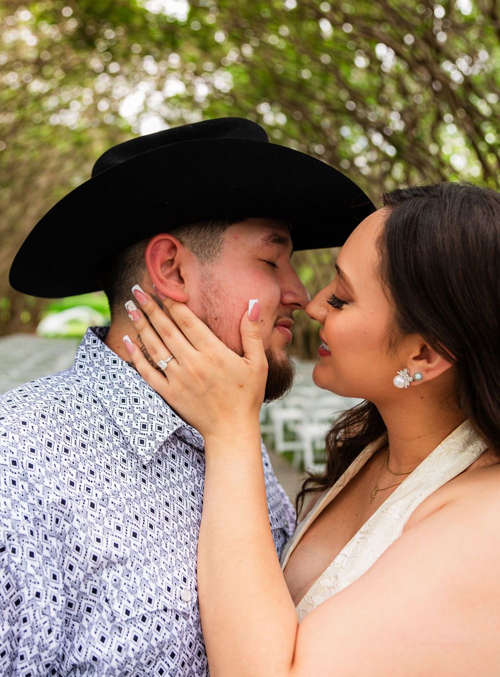 Couple sharing a romantic moment, woman in white dress touching mans face, man wearing black cowboy hat outdoors.