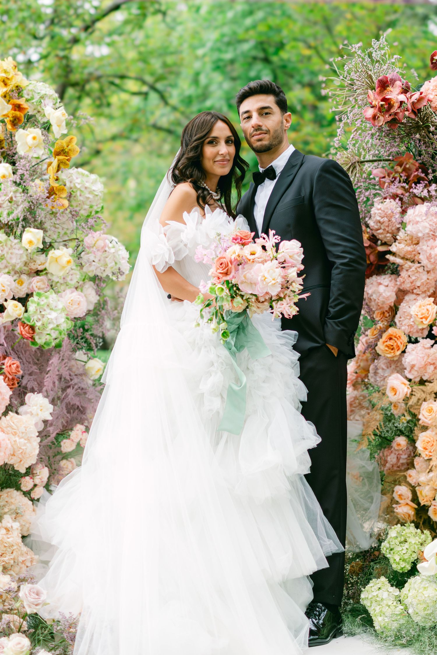 Bride in ruffled white gown with bouquet and groom in black tuxedo pose before lush floral backdrop outdoors.