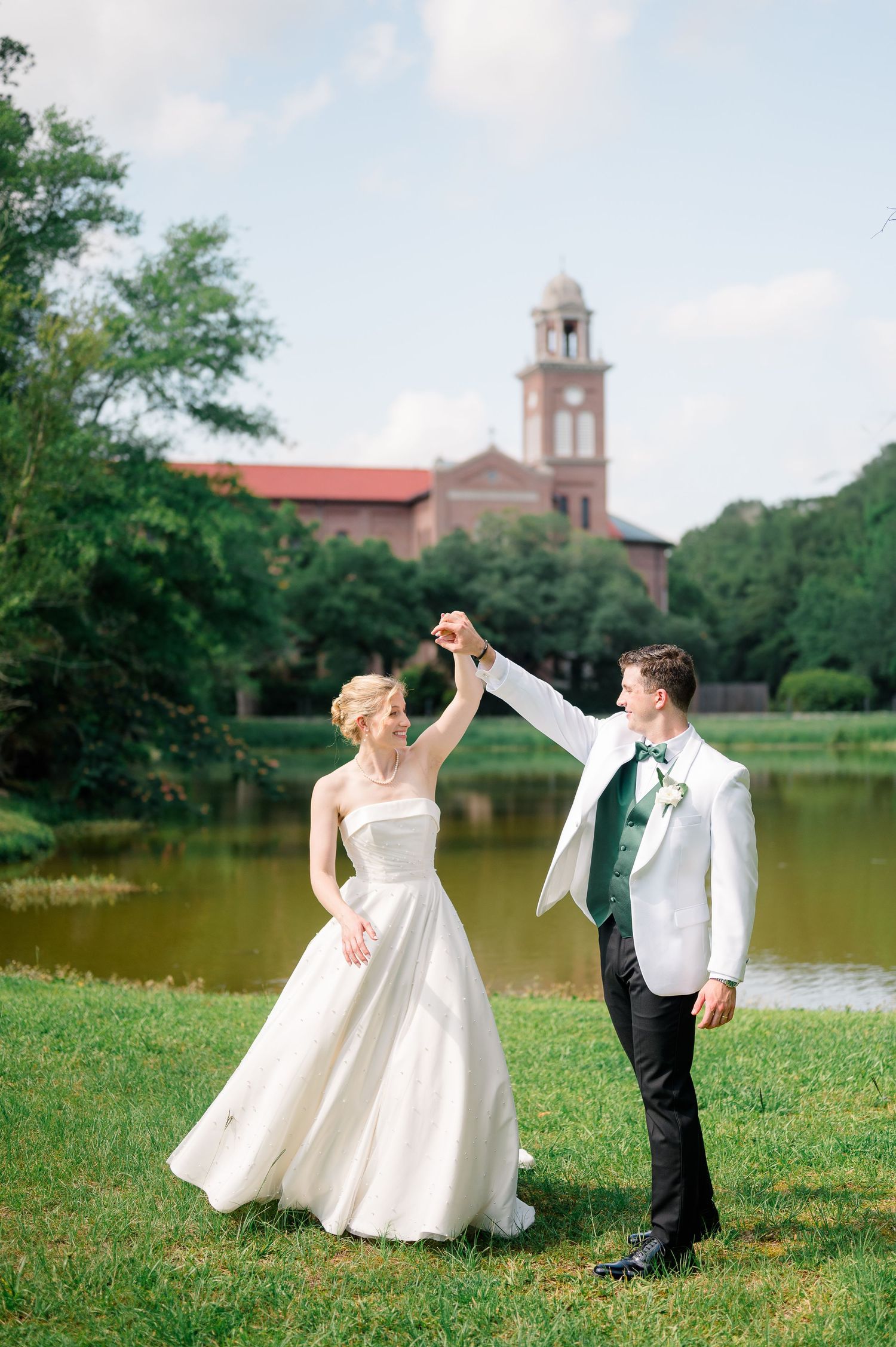 Bride and groom dancing outdoors by a lake with a historic brick church and lush green trees in the background.