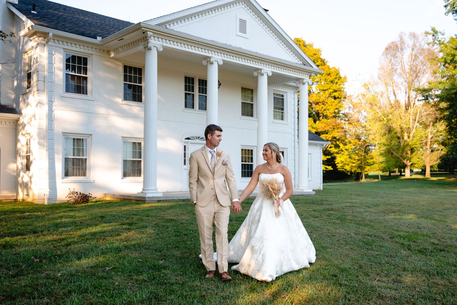 Bride and groom walk hand-in-hand on green lawn in front of elegant white colonial mansion.