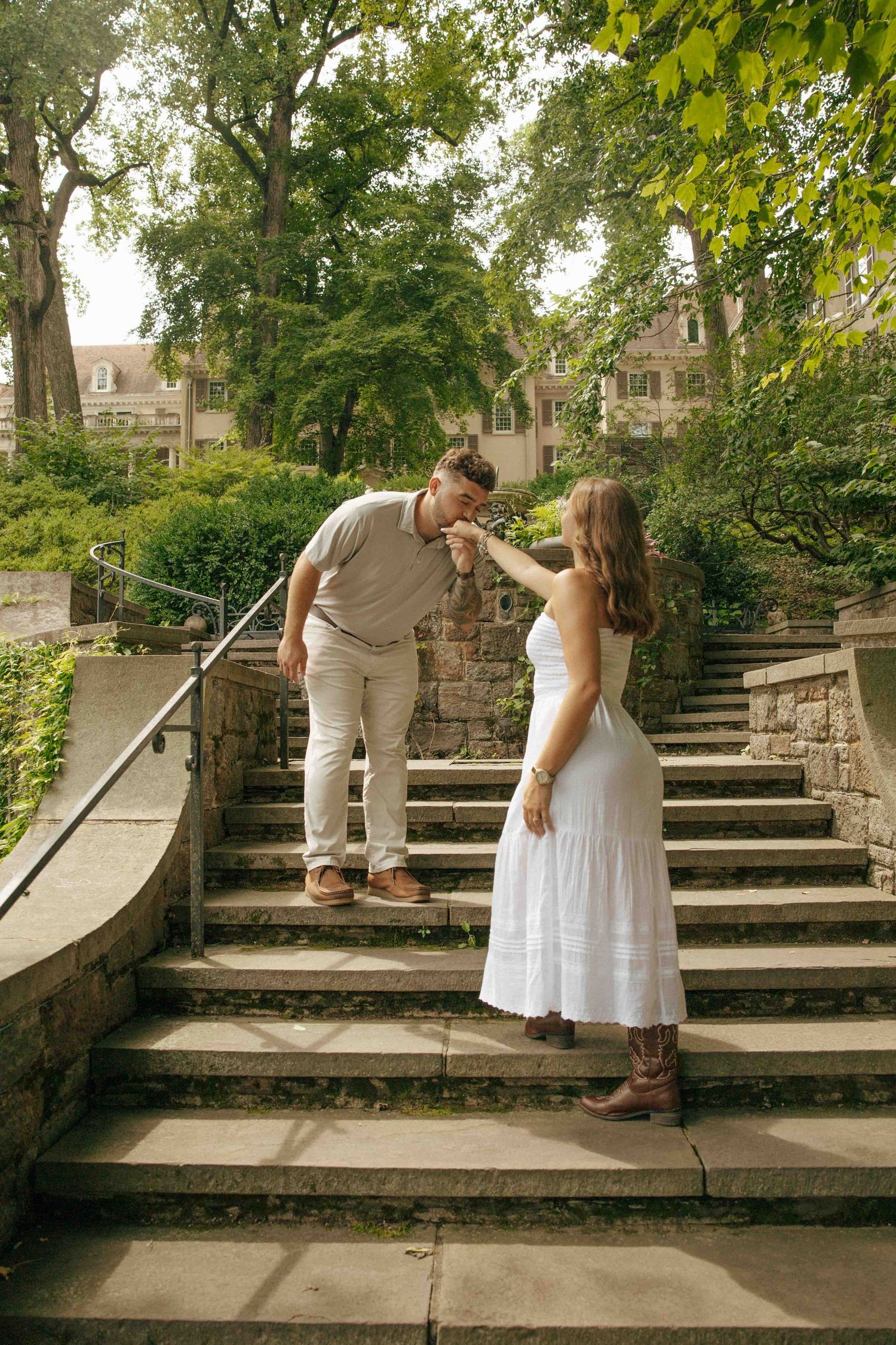 A man kisses a woman's hand on stone steps surrounded by lush greenery, as she wears a white dress and cowboy boots.