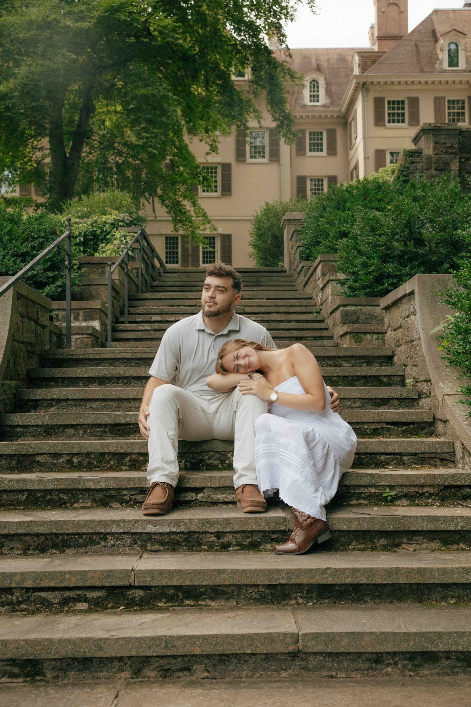 Couple sitting on stone steps outside a historic mansion, woman in white dress leaning on man in casual attire.