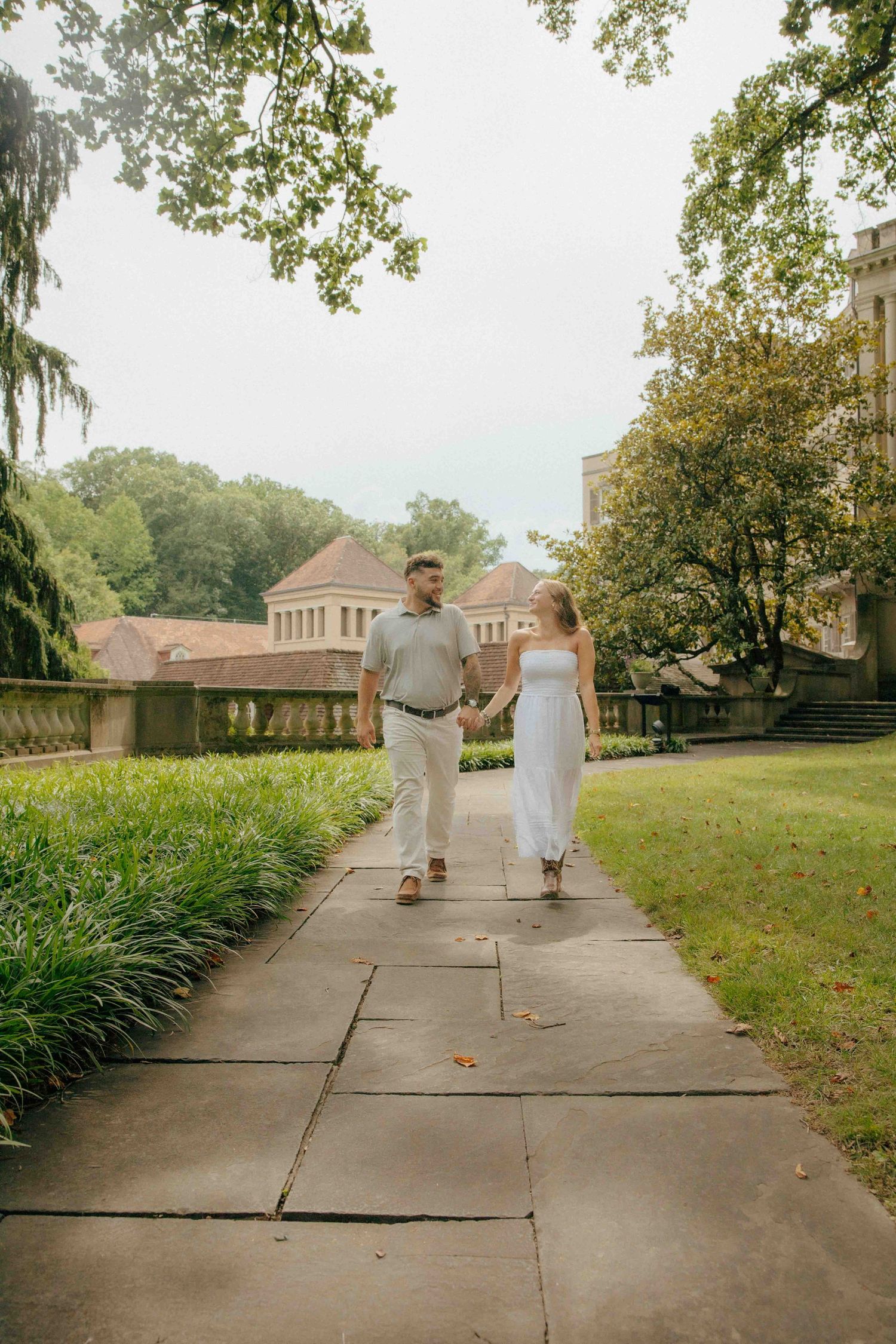 Couple holding hands walking on a stone path through lush green gardens surrounded by historic buildings.