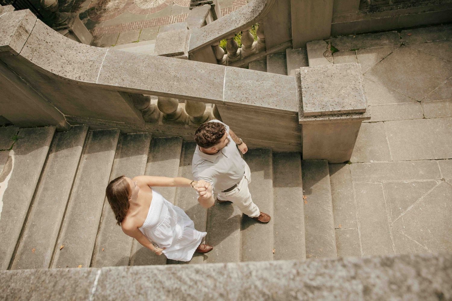 Aerial view of a couple holding hands while walking up grand stone stairs, dressed in white wedding attire.