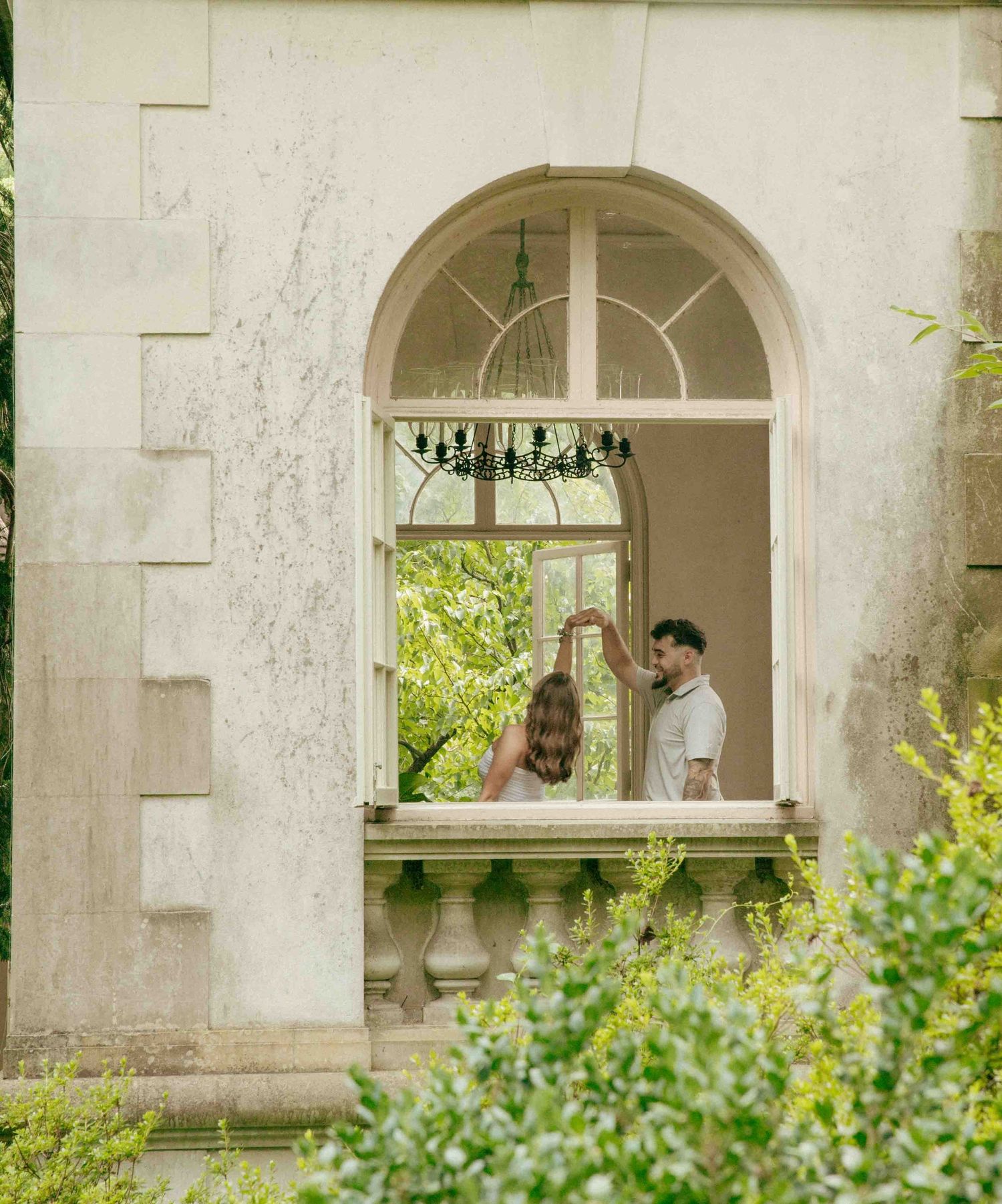 Couple dancing on a balcony framed by an elegant arched window of a stone mansion surrounded by lush greenery.