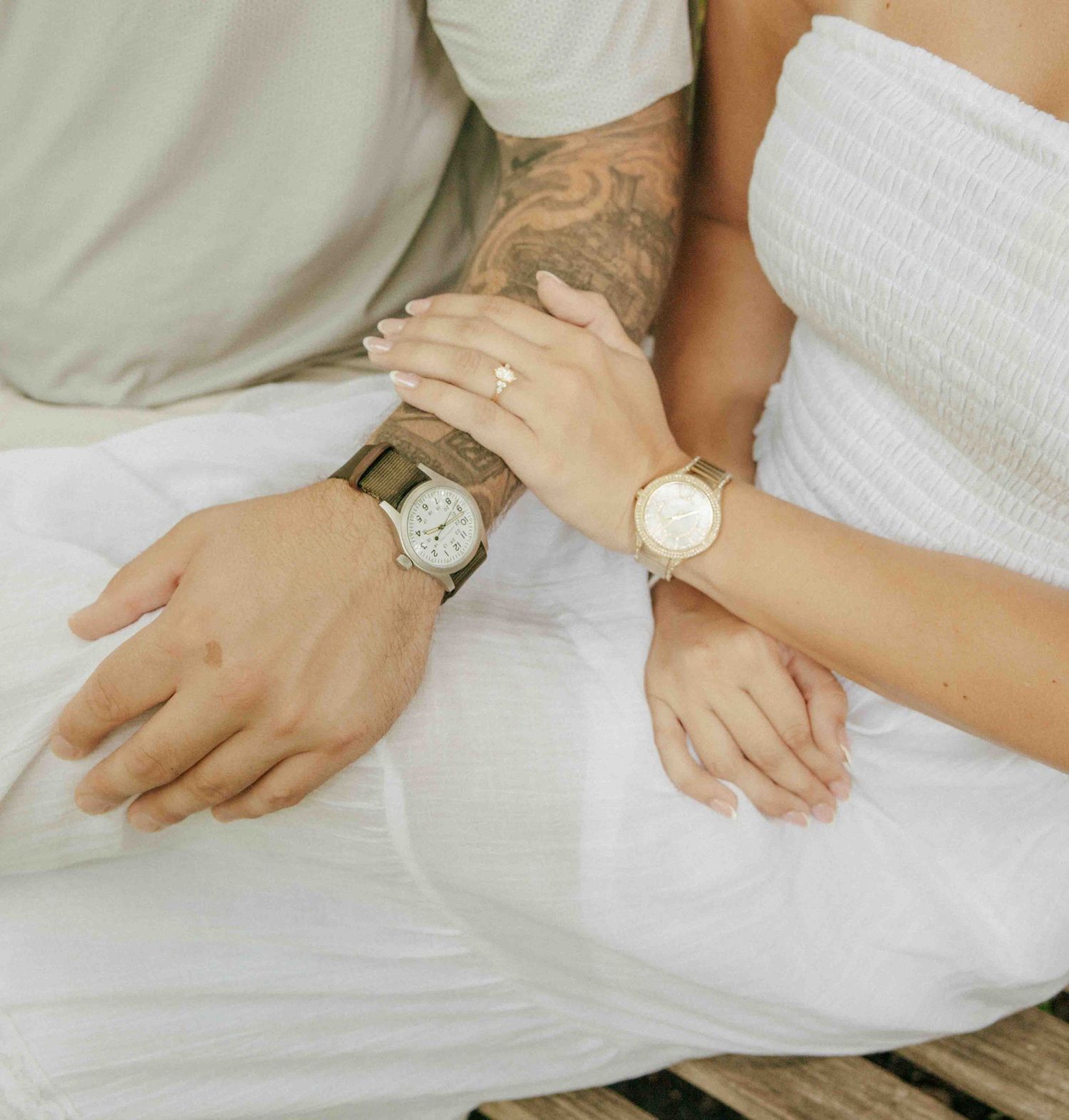 Couple wearing matching his and hers wristwatches, sitting together in white outfits, man with tattooed arm.