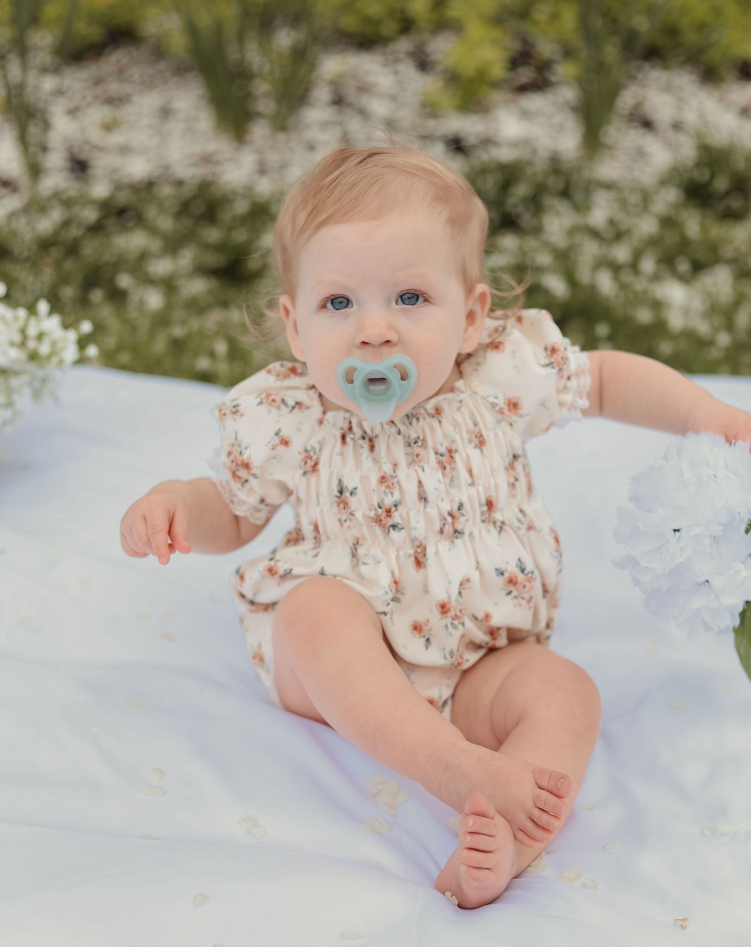 An adorable baby in a floral romper sits on a white blanket outdoors with a blue pacifier.