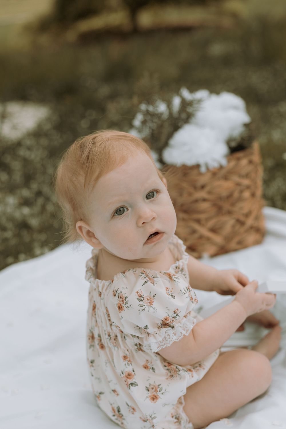A cute red-haired baby in a floral romper sits on a white blanket near a wicker basket outdoors.