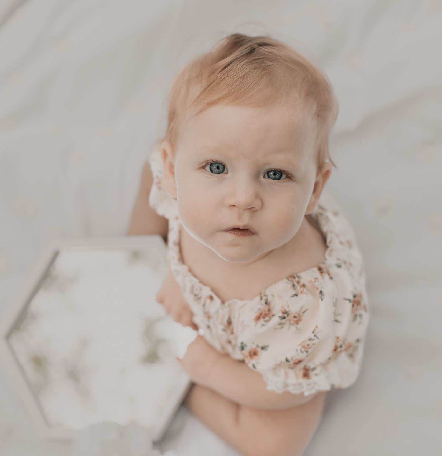 A sweet baby in a floral outfit looks up at the camera while sitting on a white surface.
