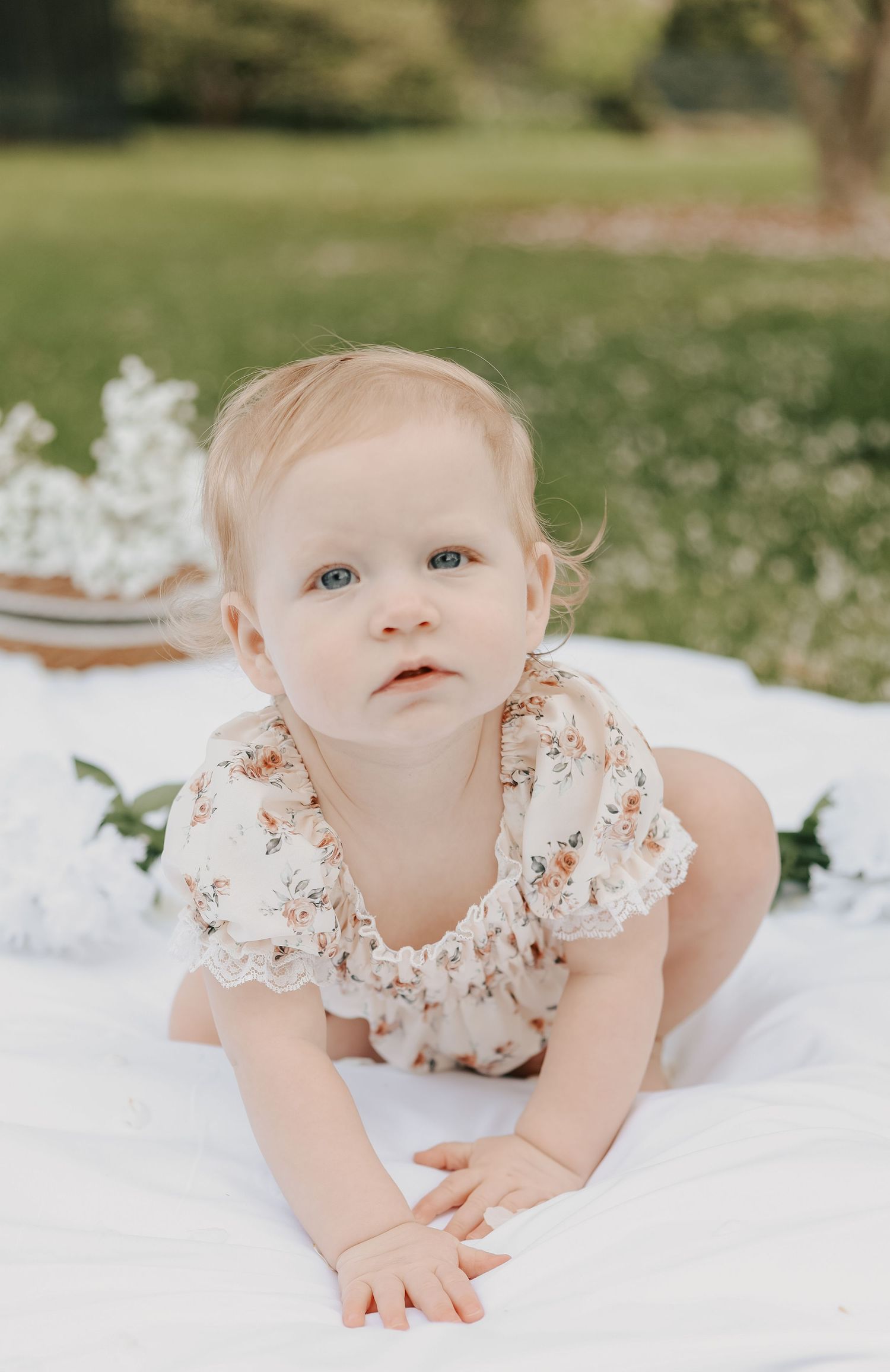 Adorable baby girl in floral romper crawling on white blanket in lush green garden.