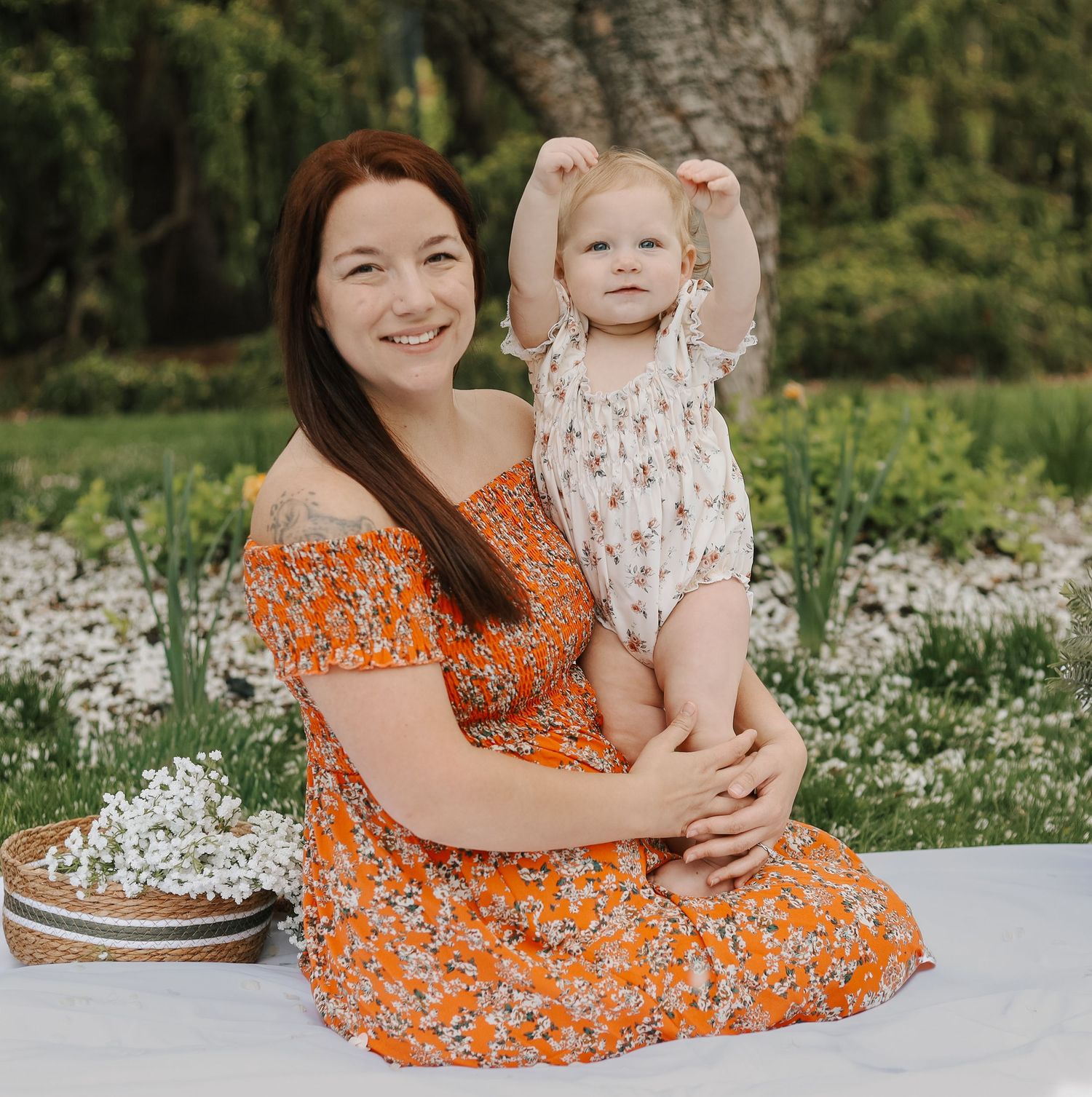 Joyful mother sitting on white blanket holding baby girl with arms raised in garden.