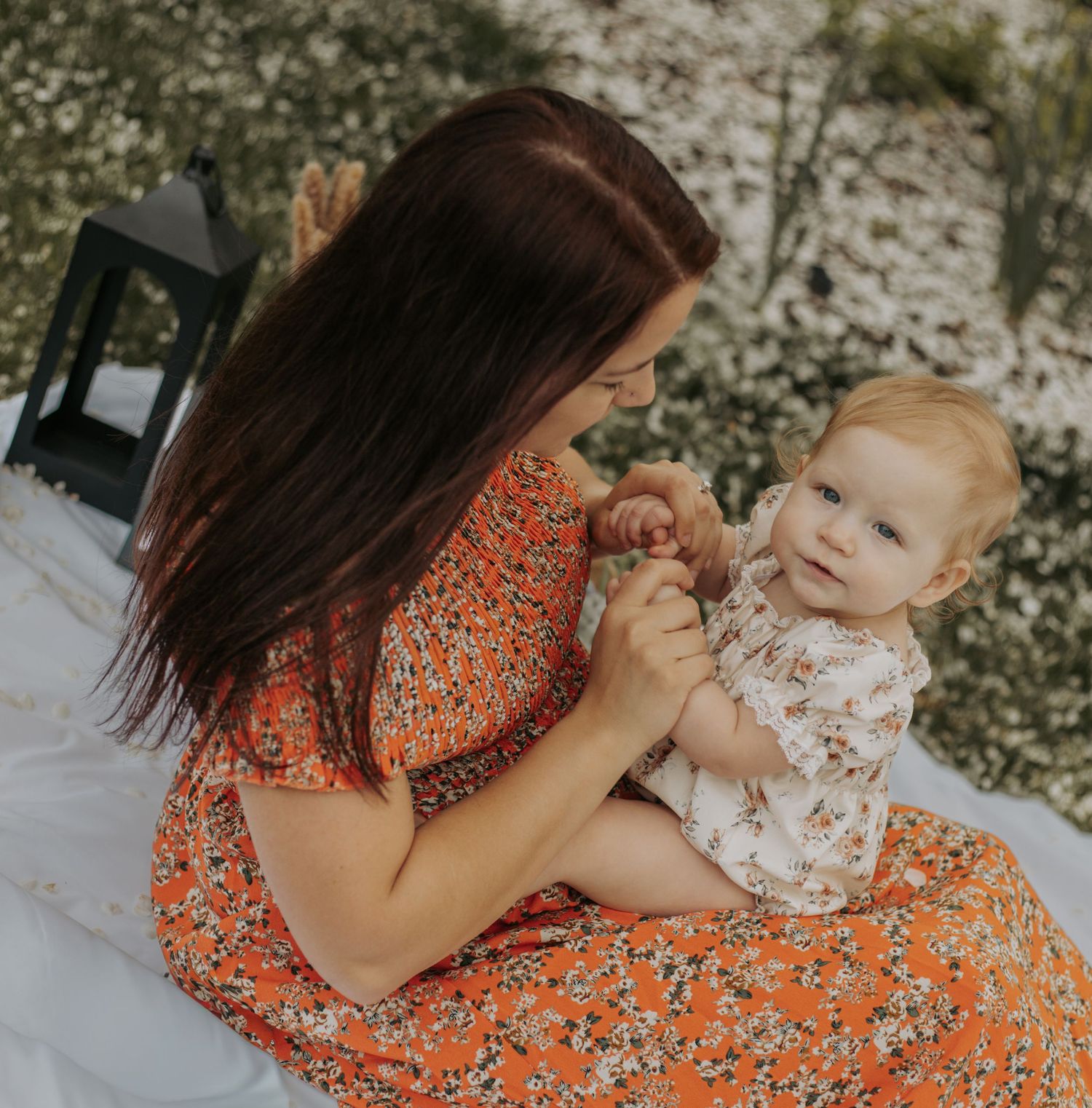 Loving mother in orange dress holding baby girls hands while sitting outdoors together.