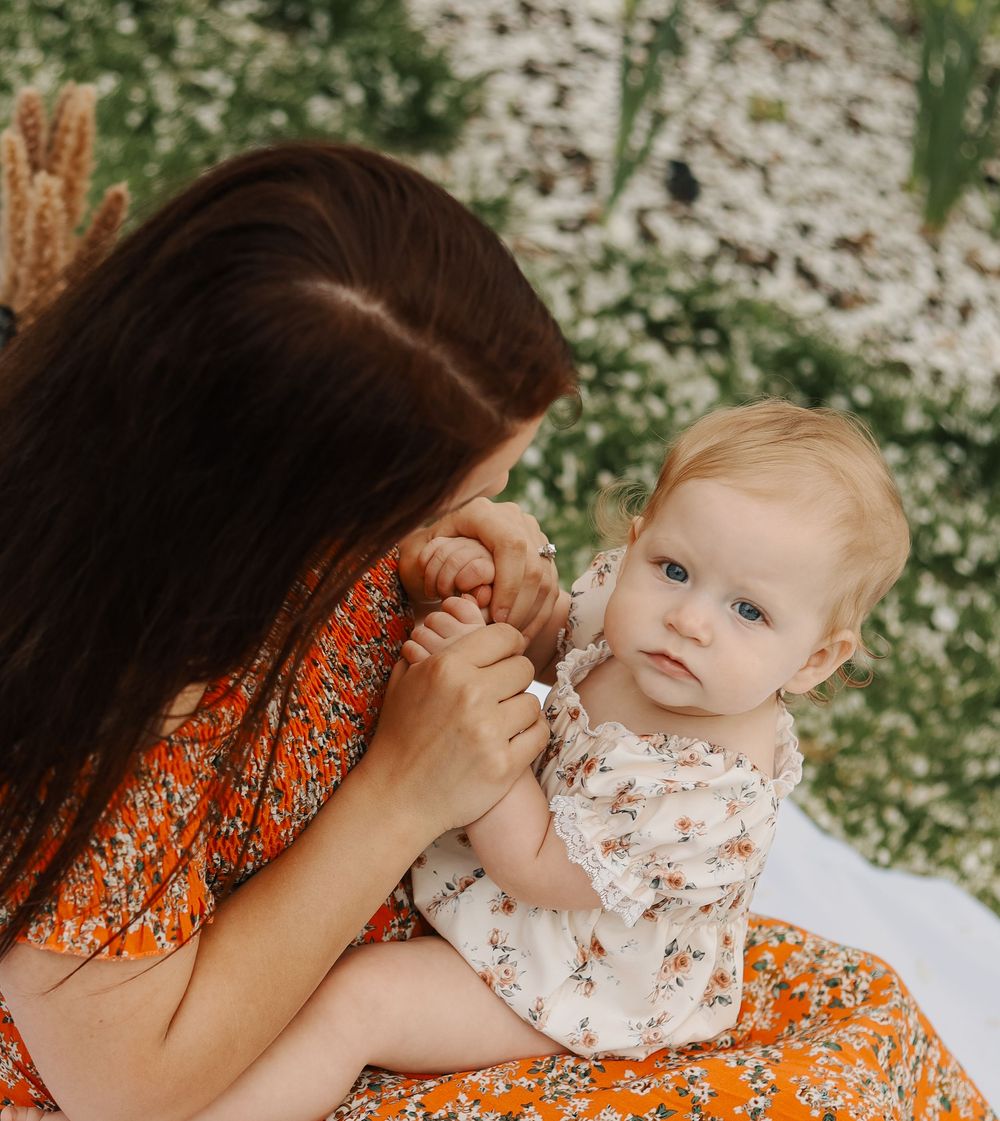 Mother in orange floral dress tenderly kissing baby girls hand in garden setting.