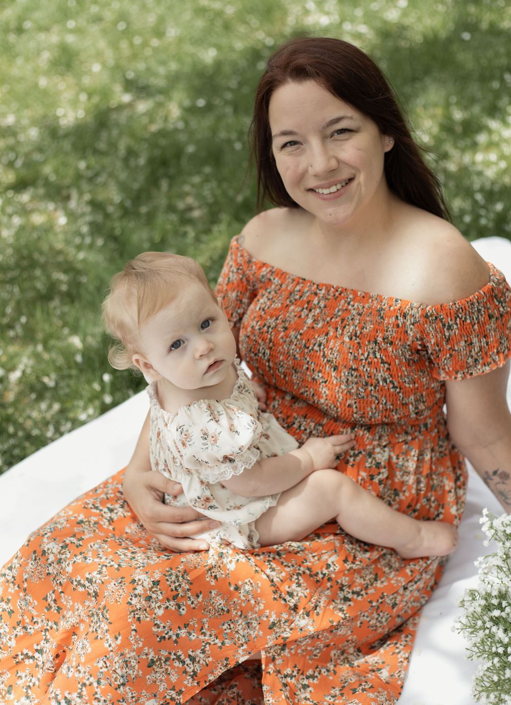 Smiling mother in off-shoulder orange dress cuddling baby girl on white blanket outdoors.