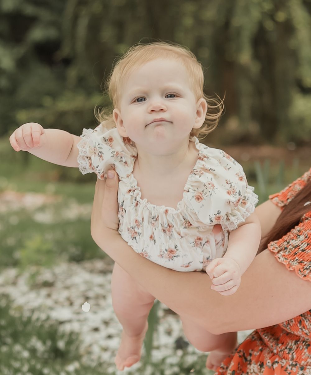 Cute baby girl in floral romper making funny face while being held in garden.