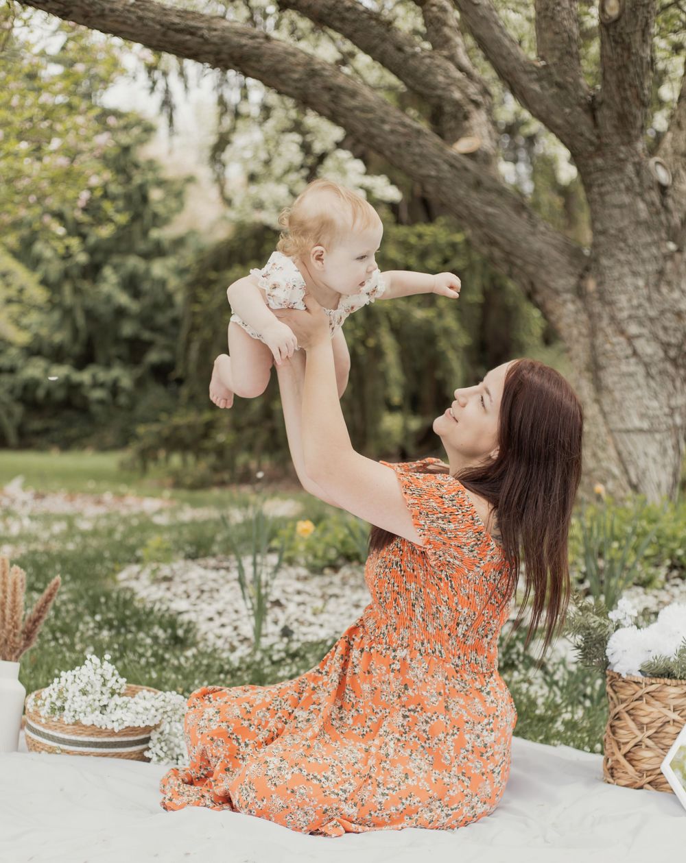 Mother in orange floral dress lifting smiling baby in the air outdoors under a tree.