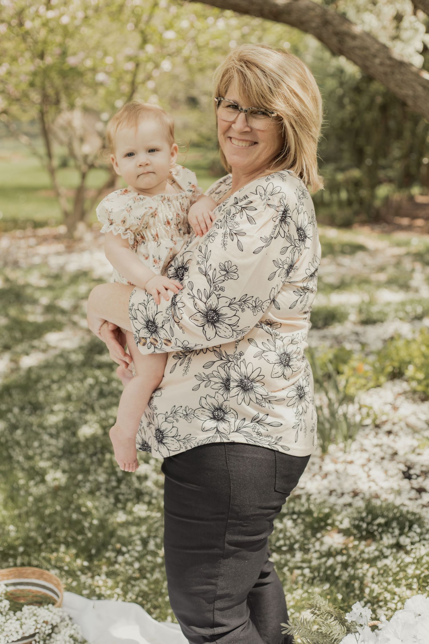 Grandmother with glasses holding baby in matching floral outfits in a garden.