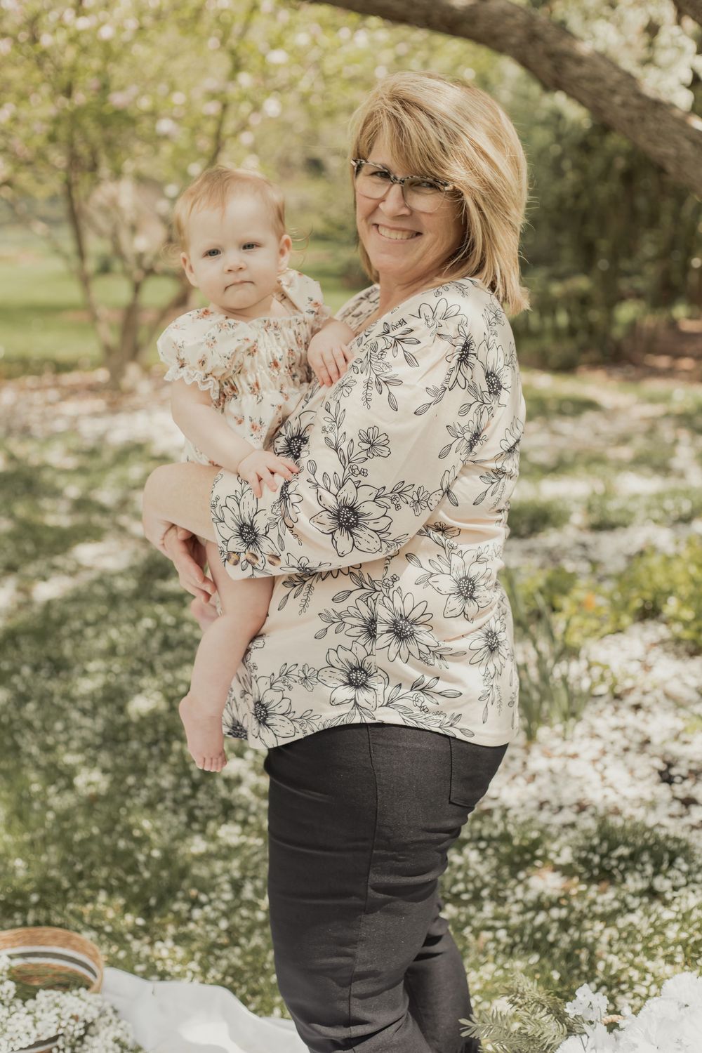 Grandmother with glasses holding baby in matching floral outfits in a garden.