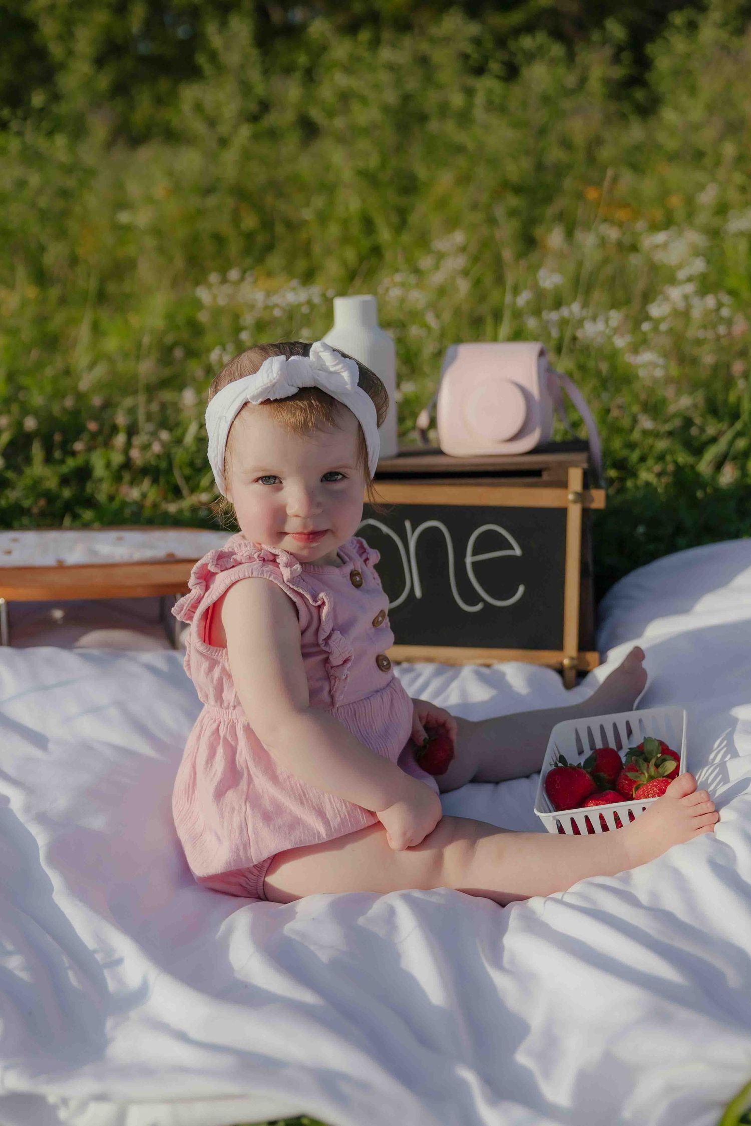 Adorable baby girl in pink romper sitting on white blanket outdoors, holding strawberries near a chalkboard sign reading one.