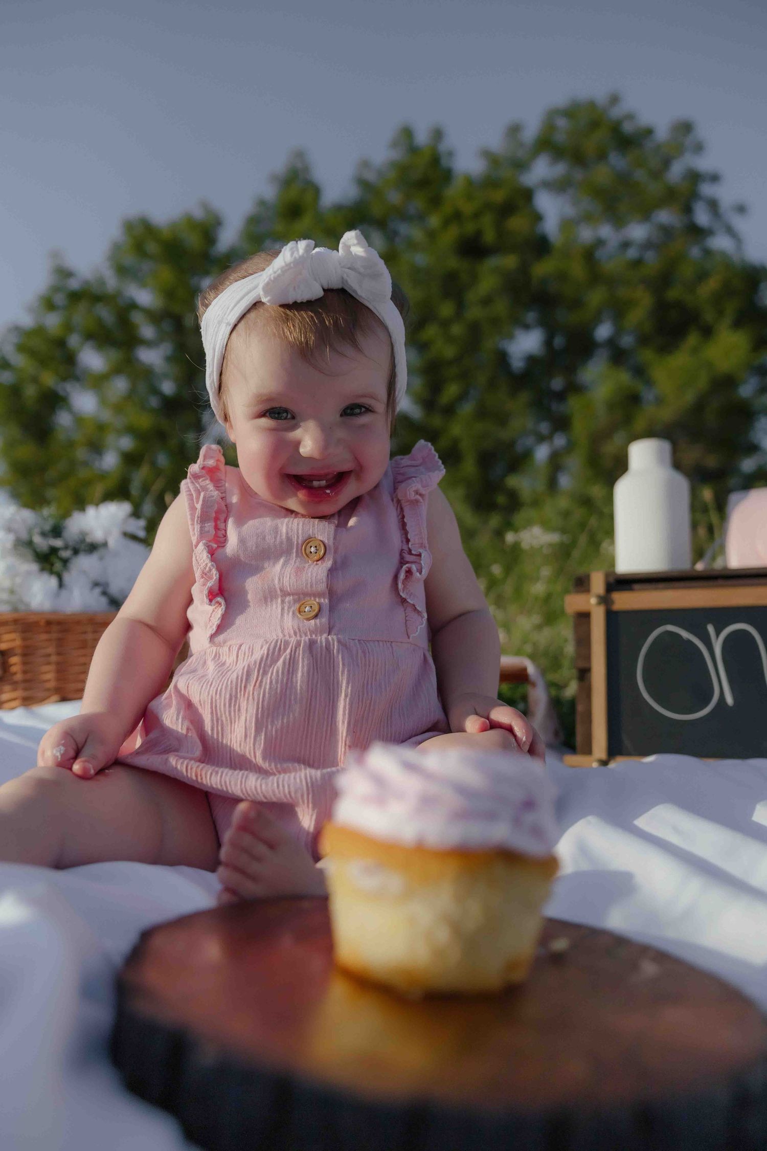 A smiling baby girl in a pink dress and white bow headband sits outdoors near a cupcake on a wood slice for her first birthday.