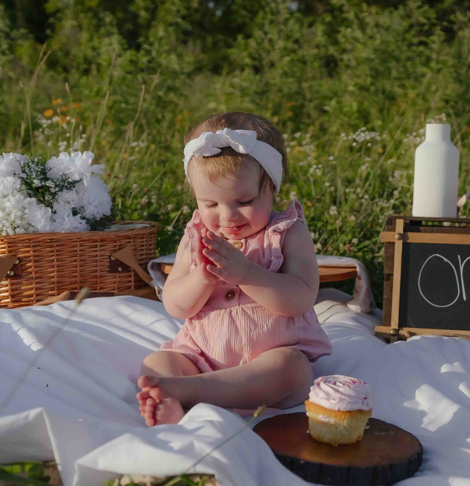 Baby girl in pink romper sits on white blanket outdoors, clapping near a cupcake, with flowers and picnic basket nearby.