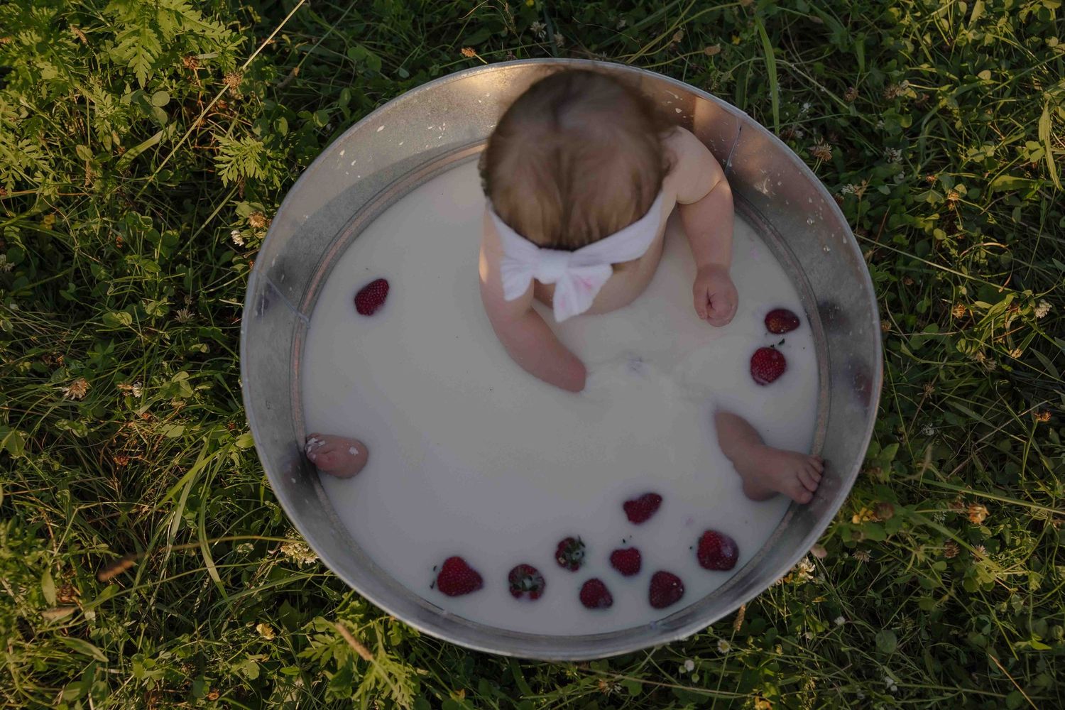 Baby sitting in a round metal tub filled with milk and fresh berries on green grass outdoors.