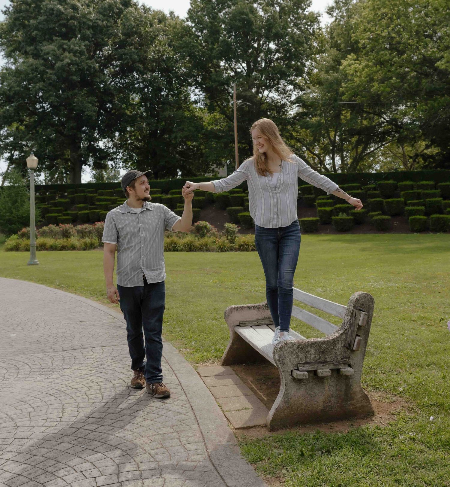 A woman stands on a park bench holding hands with a boy walking beside her in a lush green park.