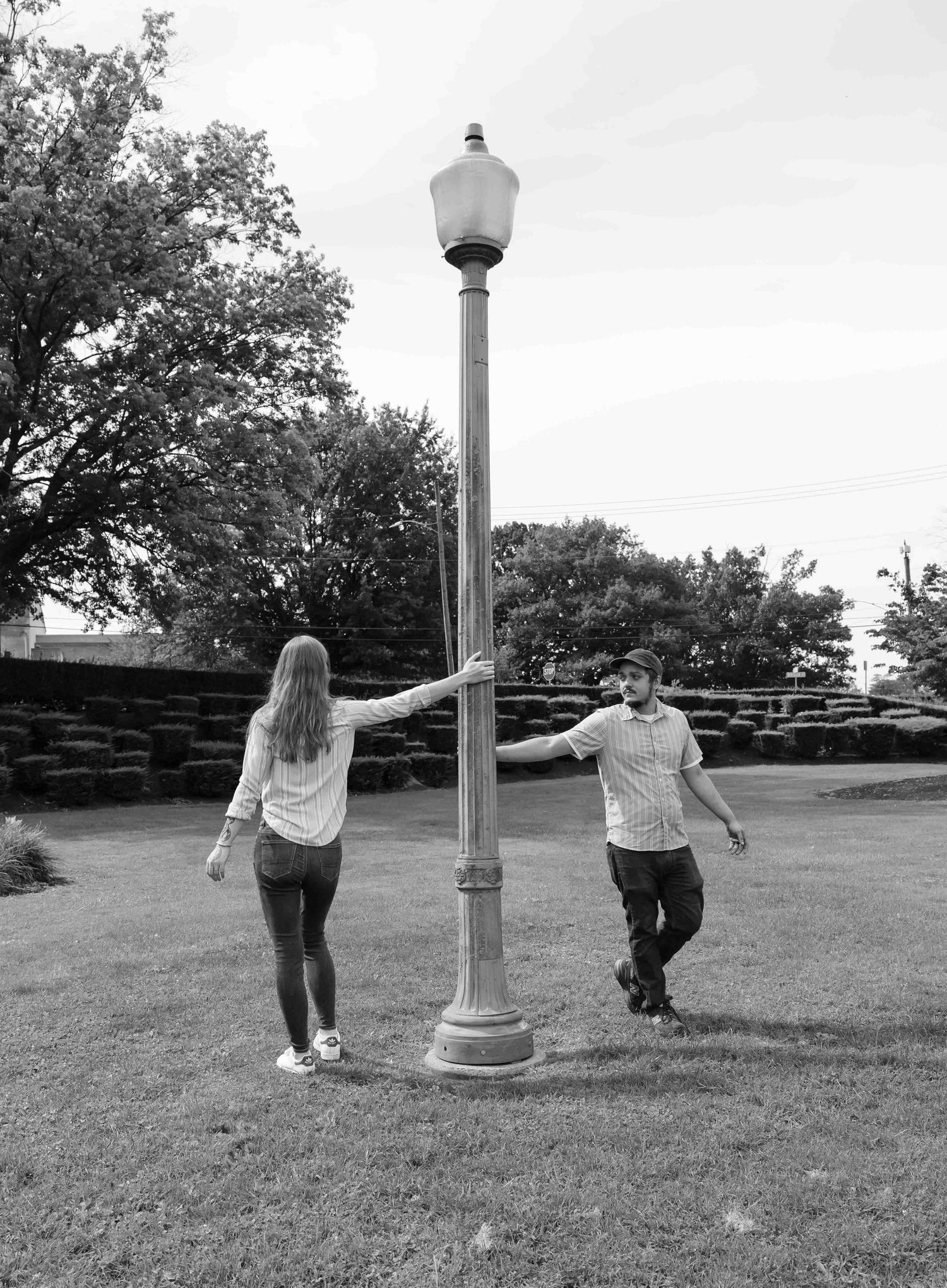 Black and white photo of a couple holding hands around a vintage lamp post in a park.