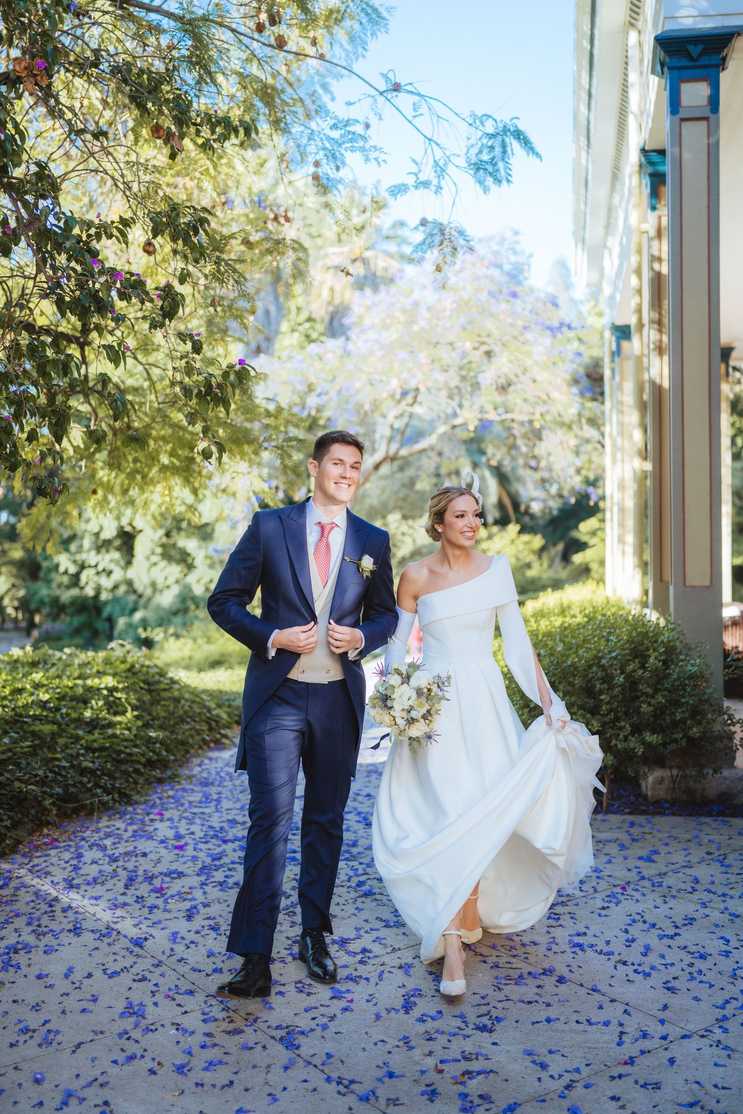Bride and groom walk together on a petal-covered path surrounded by lush gardens on their wedding day.