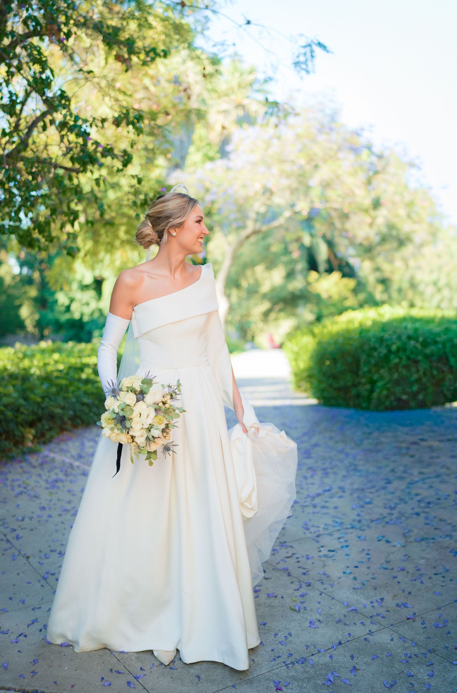 Bride in elegant one-shoulder white gown with gloves holds bouquet on a tree-lined garden path.