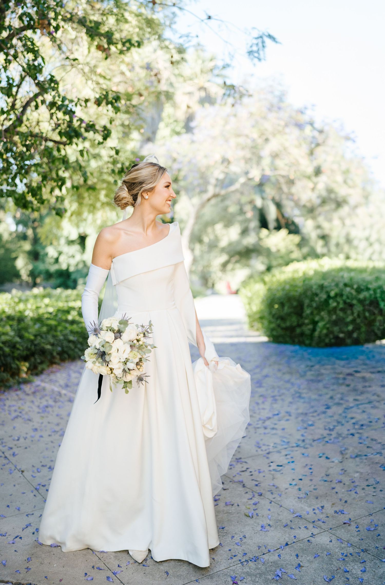 Bride in elegant one-shoulder white gown with gloves holding floral bouquet in a lush garden setting.