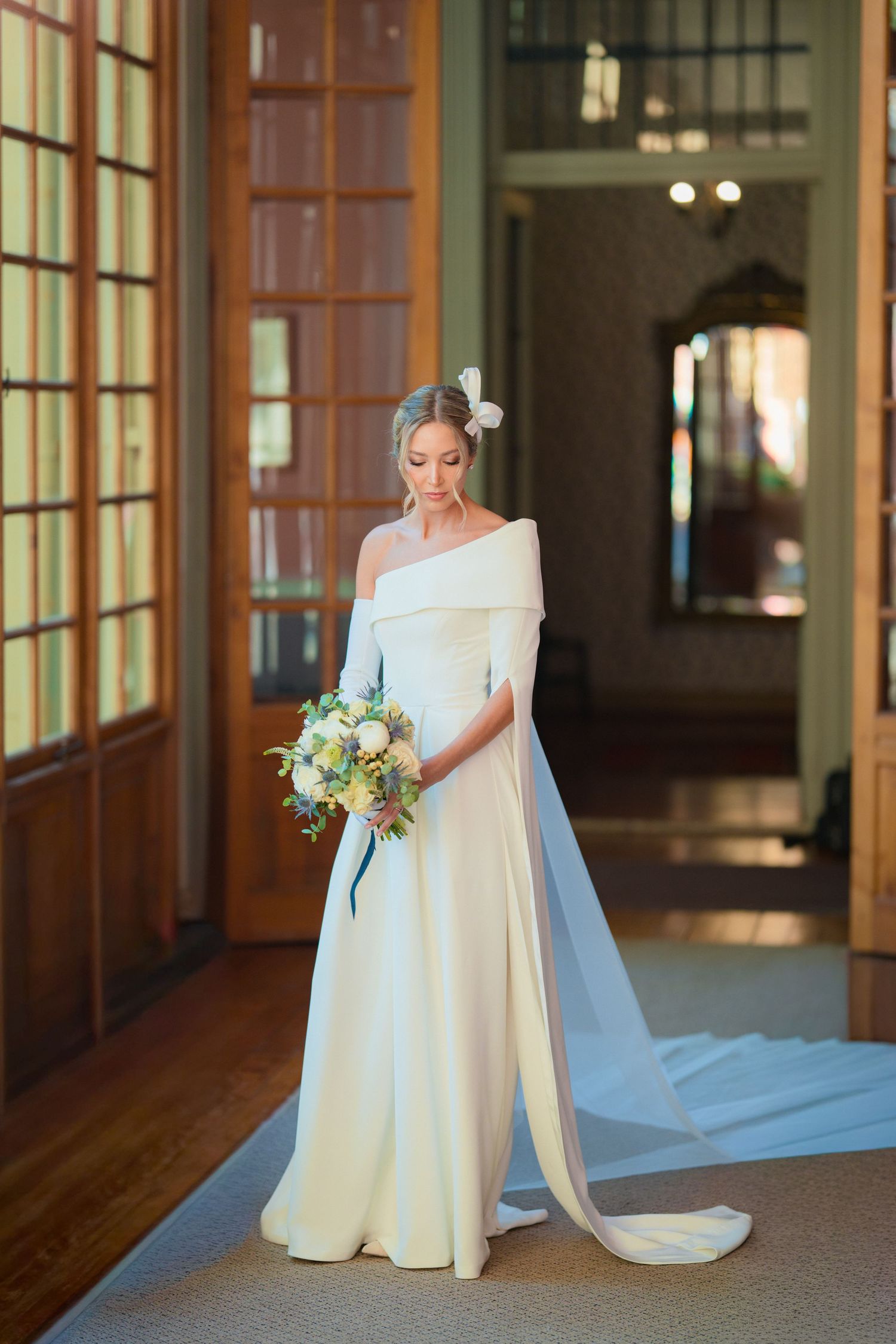 Bride in elegant off-shoulder white gown with cape train holds floral bouquet in a grand wood-paneled hall.