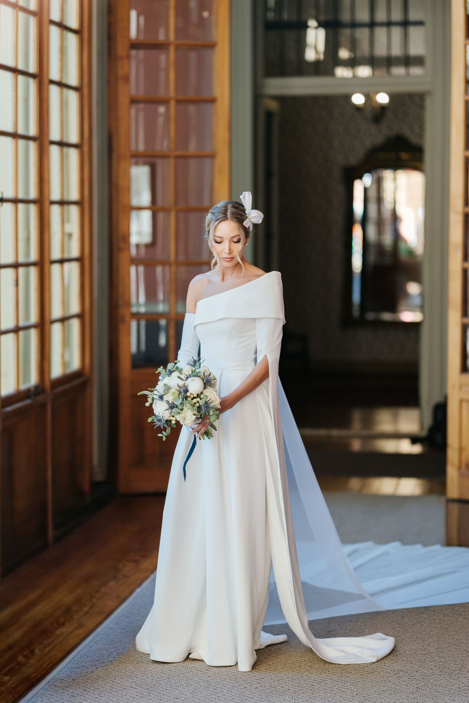 Elegant bride in a white off-shoulder gown with cape train holding a floral bouquet in a sunlit venue.