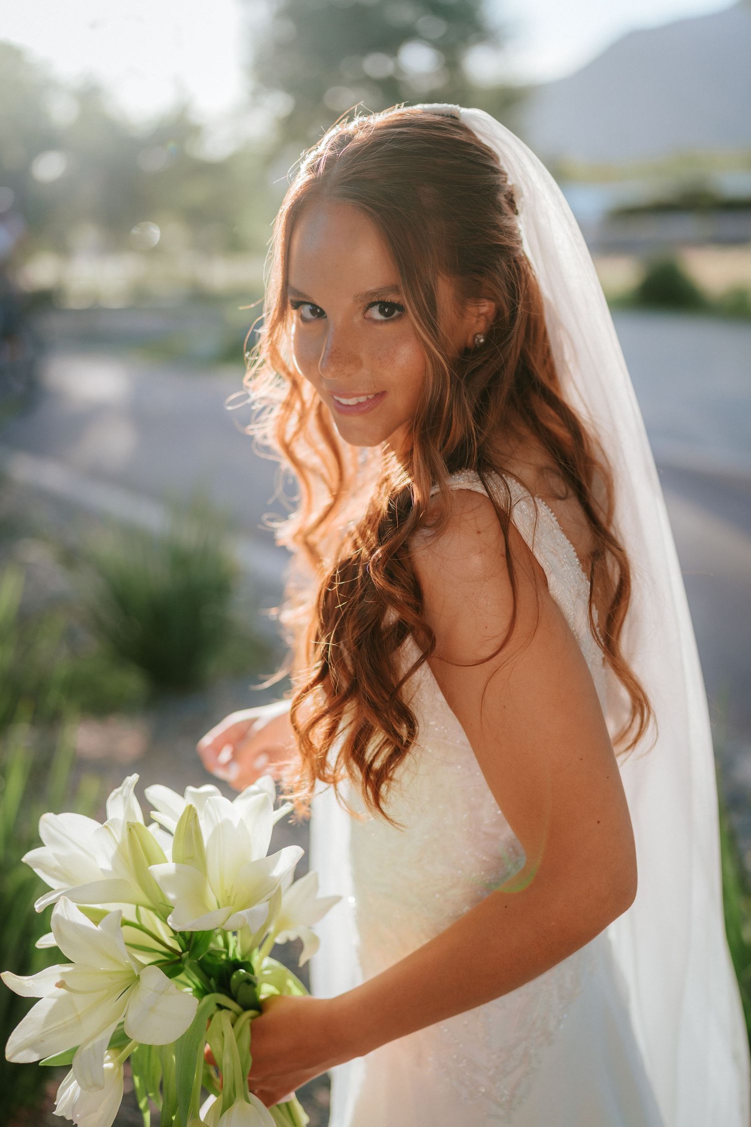 A radiant bride with long auburn hair and white veil holds a bouquet of white lilies in a sunlit outdoor setting.
