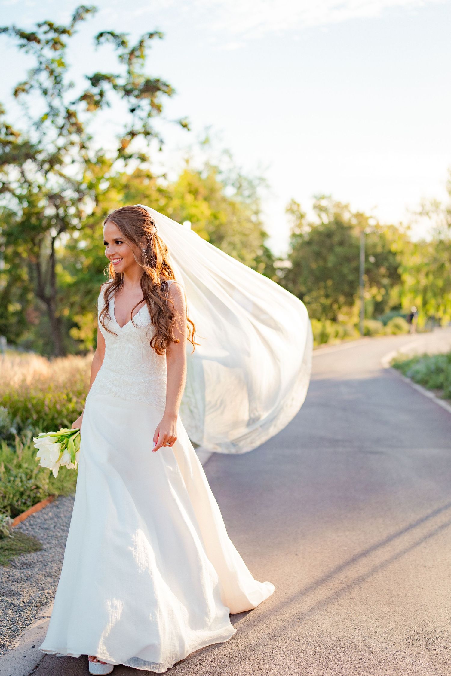 Smiling bride in white gown and flowing veil walks down a sunlit outdoor path holding a bouquet.