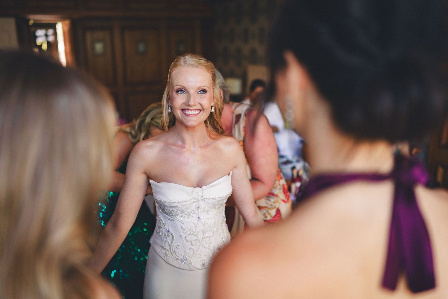 Smiling bride in a white strapless gown holding hands with guests at her wedding reception indoors.