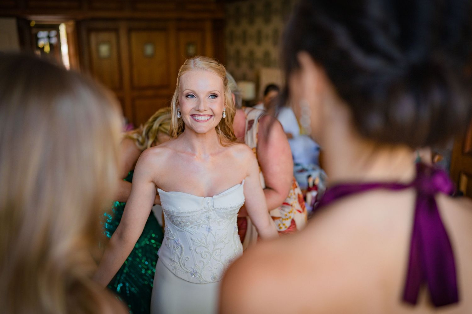 Smiling bride in strapless white gown holding hands with bridesmaids in colorful dresses at elegant wedding venue.