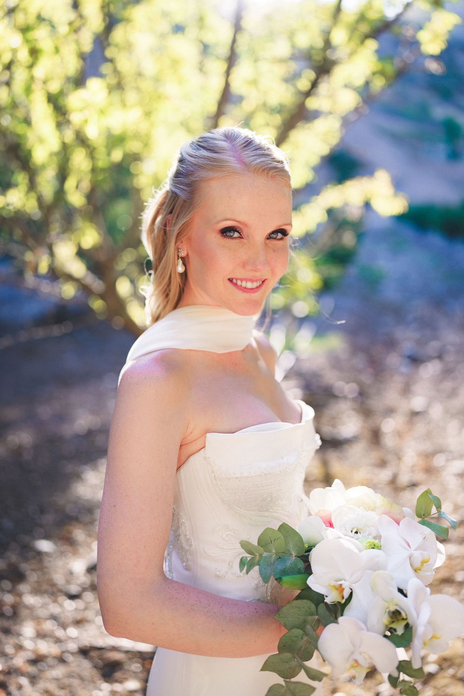 Smiling bride in strapless white gown holding white orchid bouquet outdoors in sunlit garden setting.