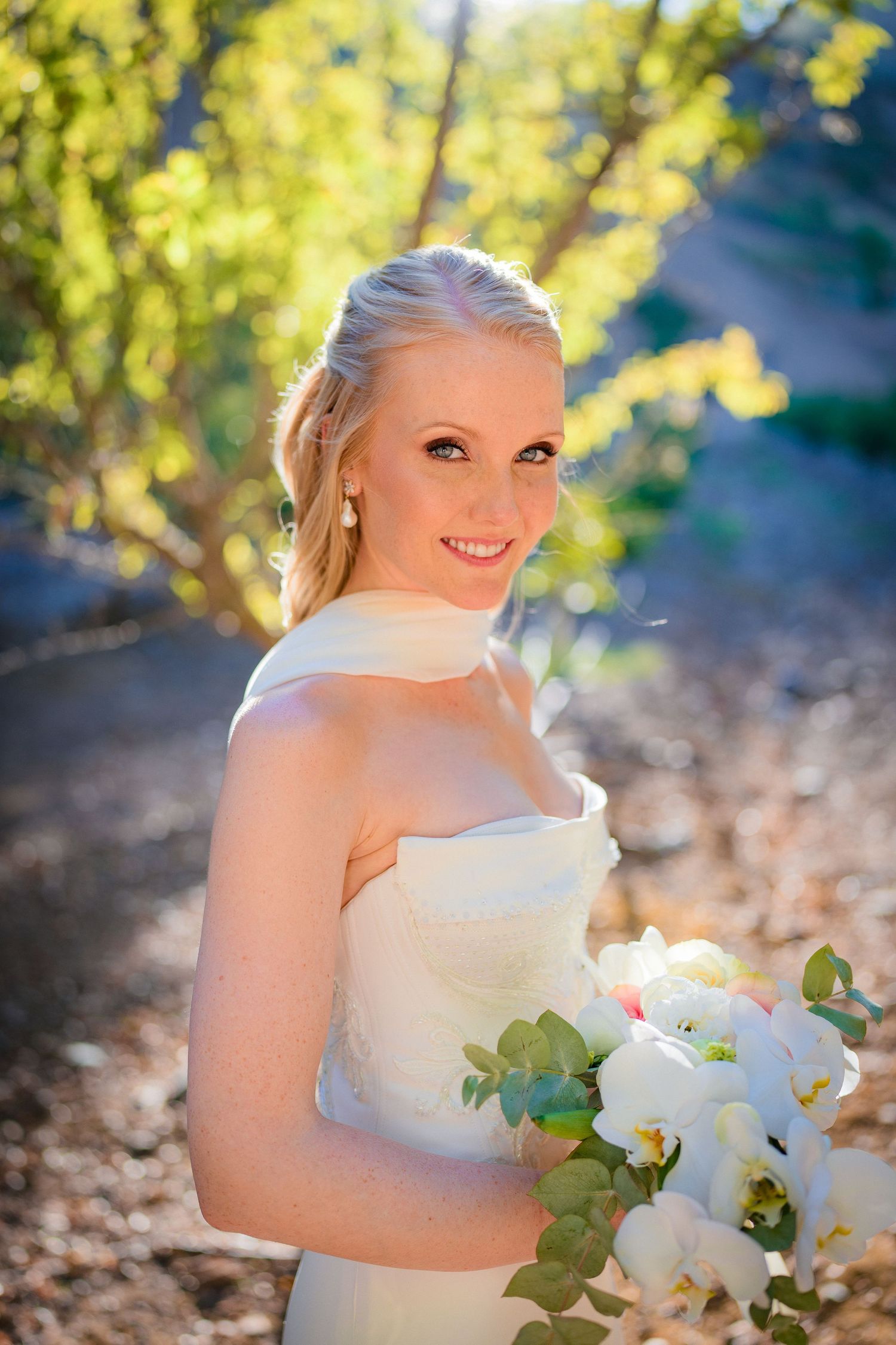 Smiling blonde bride in strapless white gown holding white floral bouquet outdoors in sunlit garden setting.