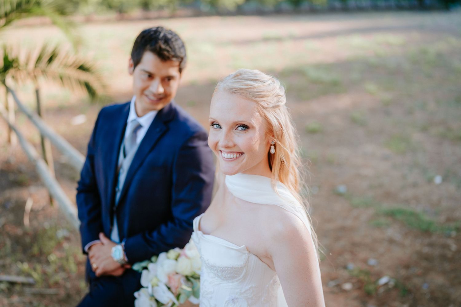 Smiling bride in strapless white gown holding bouquet with groom in navy suit standing outdoors at wedding.
