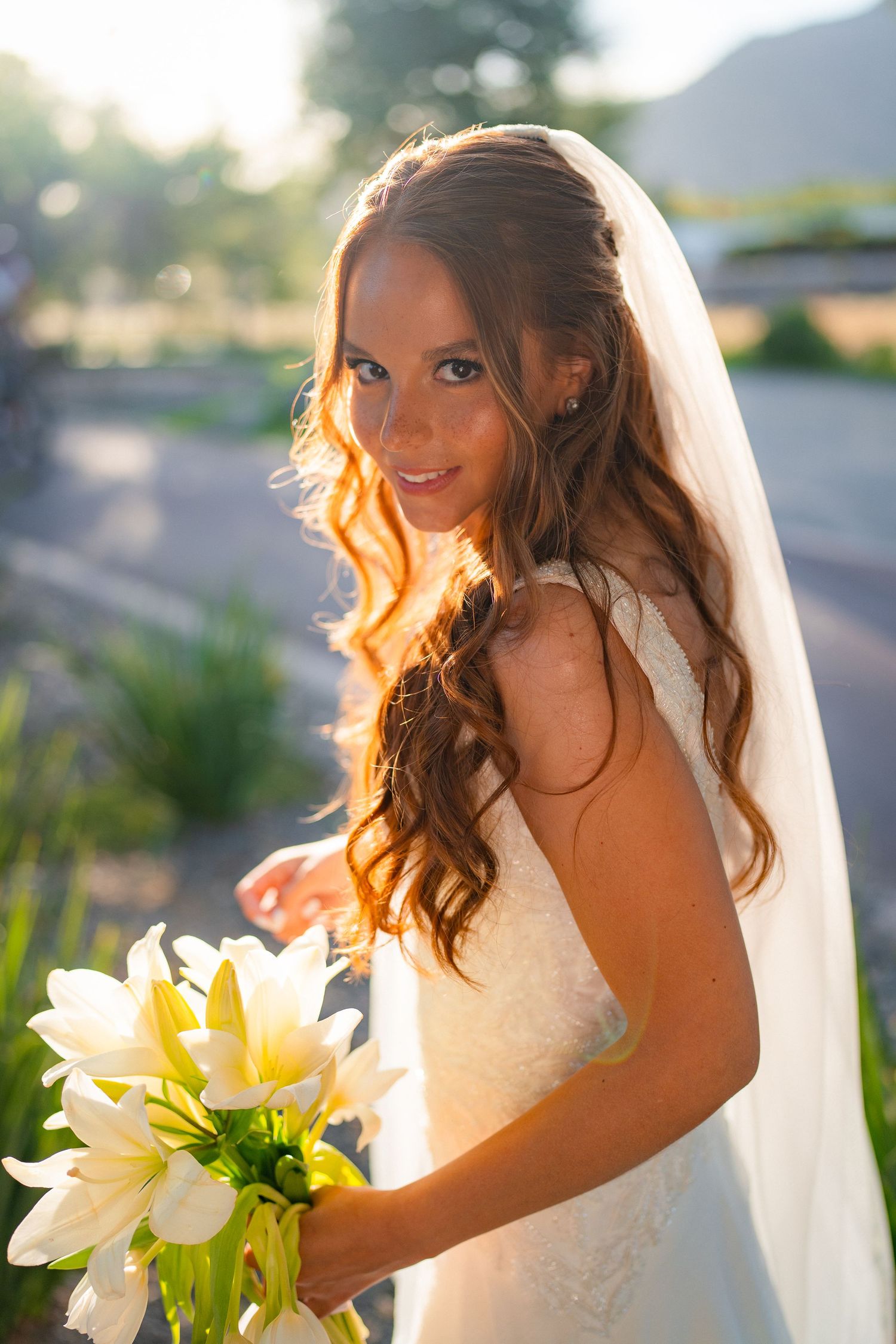 Radiant bride in white lace gown and veil holds yellow lily bouquet, glowing in warm golden sunlight outdoors.