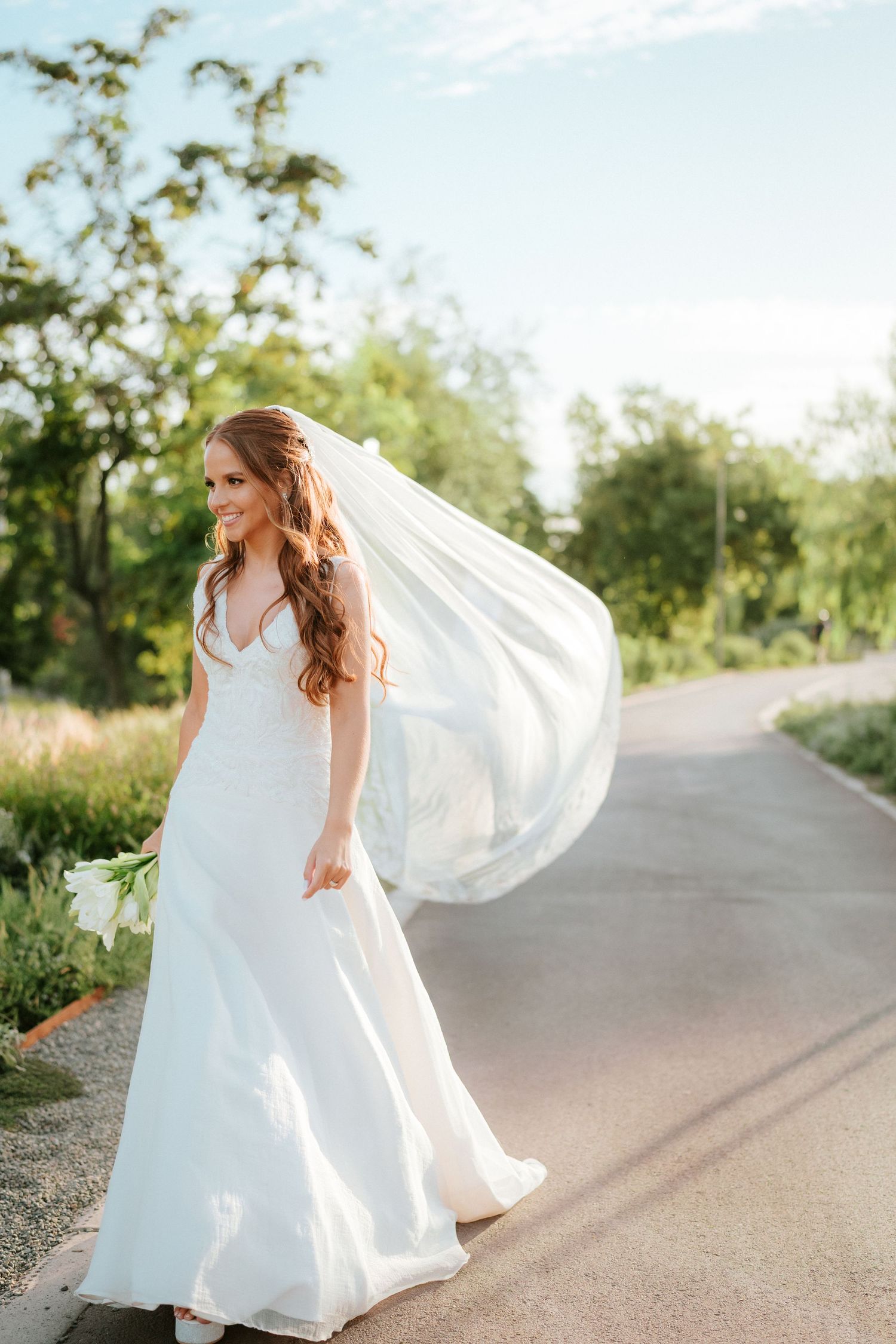 Bride in white gown with flowing veil walks on a sunlit path surrounded by lush green trees on her wedding day.