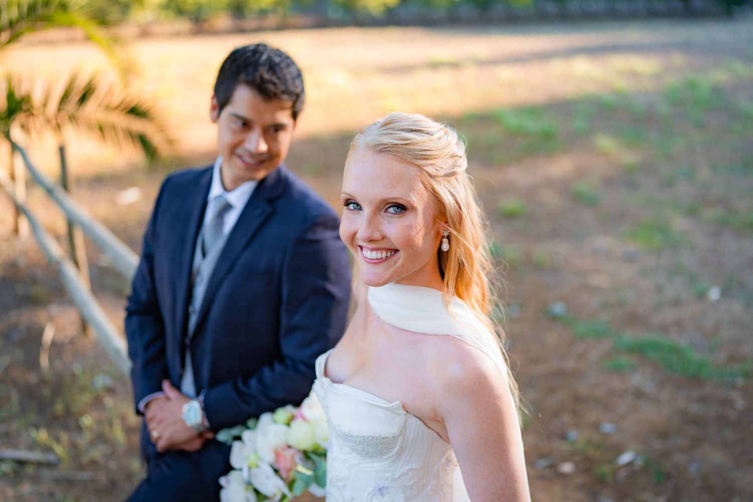 Smiling blonde bride in white strapless gown holds bouquet outdoors with groom in navy suit behind her.