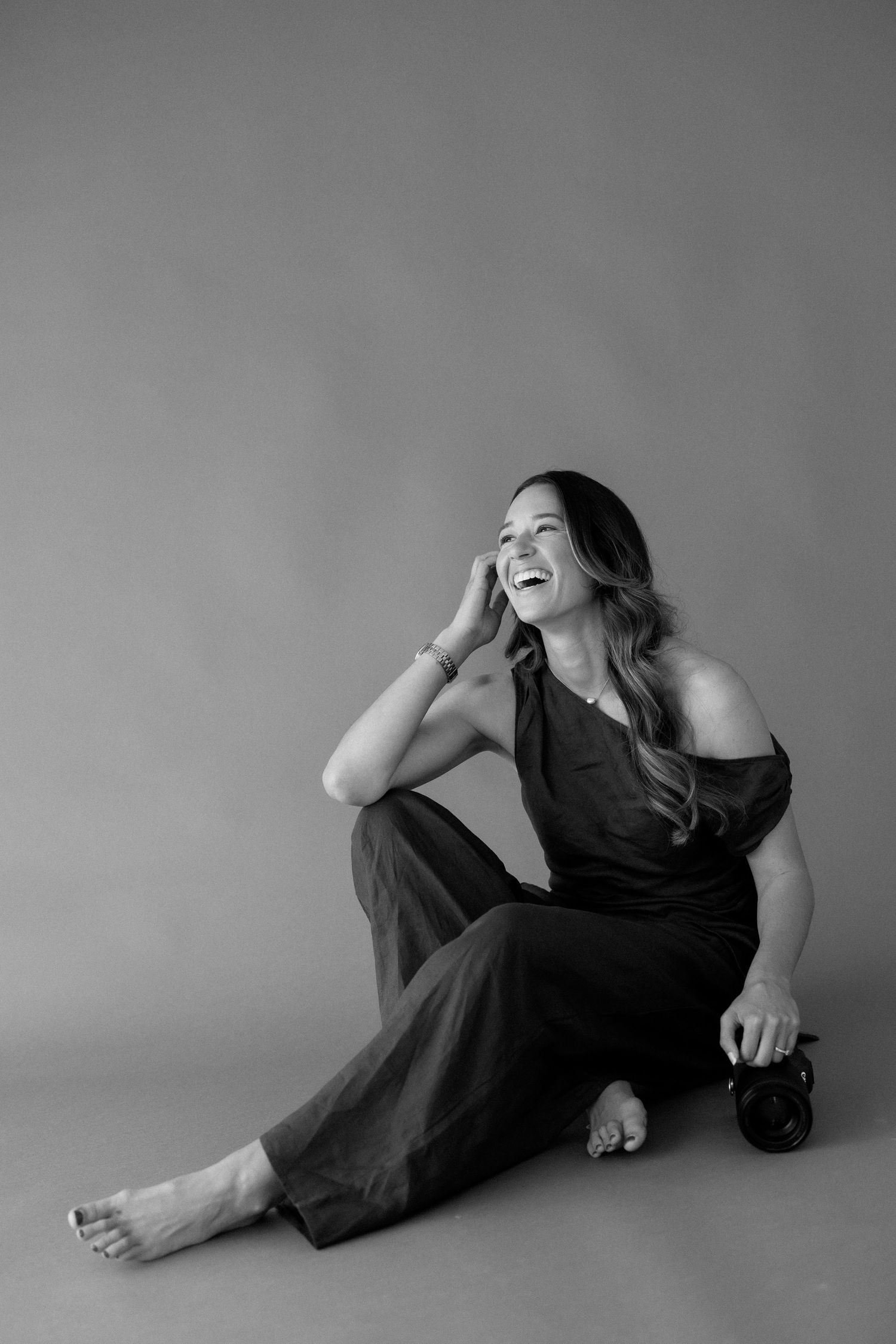 Woman in black blazer and jeans sitting on wooden chair, smiling against beige background.